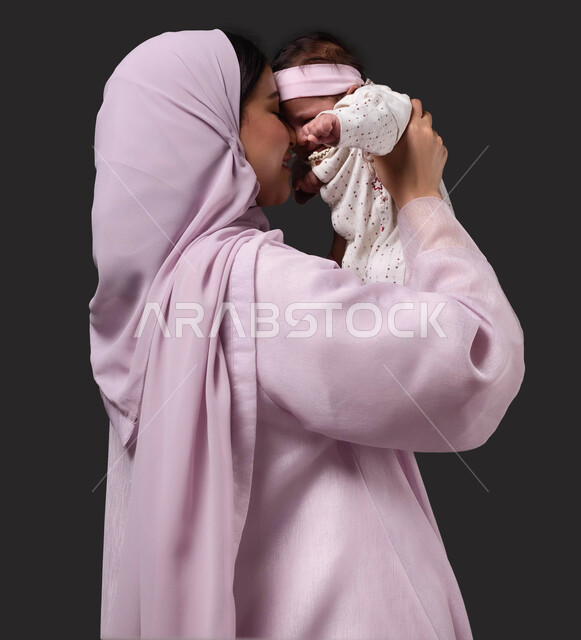 ملاعبة الأطفال بعطف واهتمام، حنان الأمهات و تحمل المسؤولية والعناية بالأطفال، قضاء الوقت باللعب مع الأبناء بإيماءات السعادة والسرور،  بورتريه من الجانب لأم عربية خليجية سعودية تبتسم ترتدي العباءة تحمل طفلتها الرضيعة وتقوم بمداعبتها بحب وحنان, خلفية سوداء