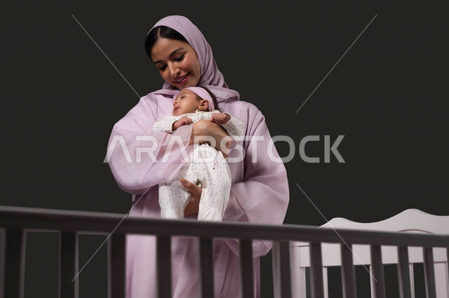 الشعور بالأمان والاطمئنان، مفهوم حنان الأمهات و تحمل المسؤولية والعناية بالأطفال، تعزيز رابط الأمومة بين الأم وصغيرتها، إيماءات السعادة والسرور، بورتريه مقرب لسيدة عربية خليجية سعودية تنظر إلى الكاميرا تحمل طفلتها الرضيعة بحب، خلفية سوداء.