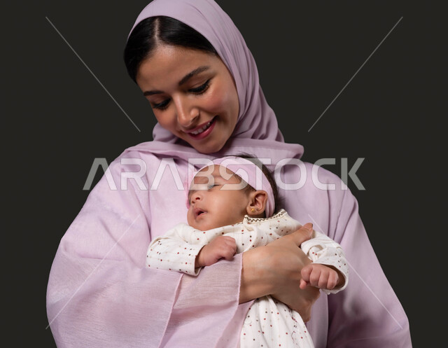 تعزيز رابط الأمومة بين الأم وصغيرتها، إيماءات السعادة والسرور، مفهوم حنان الأمهات و تحمل المسؤولية والعناية بالأطفال، بورتريه مقرب لسيدة عربية خليجية سعودية تحمل طفلتها الرضيعة تنظر إليها بحب، الشعور بالأمان والاطمئنان، خلفية سوداء.