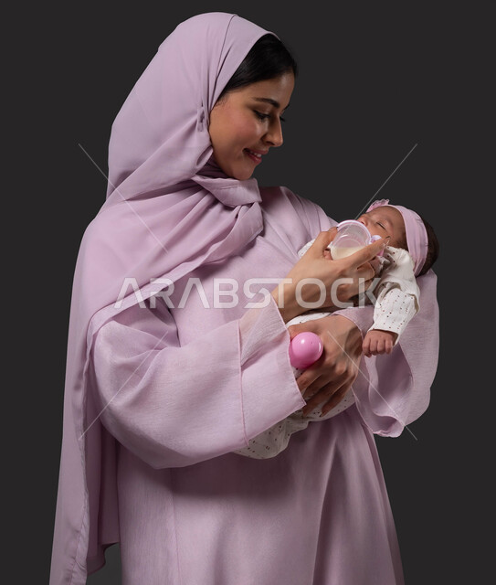 Drinking milk from a feeding bottle, caring for and paying attention to the health and development of children, the concept of motherhood and artificial feeding, a close-up portrait of a smiling Saudi Arabian Gulf mother holding her little girl, eating healthy food and meals, spending enjoyable times with children, black background