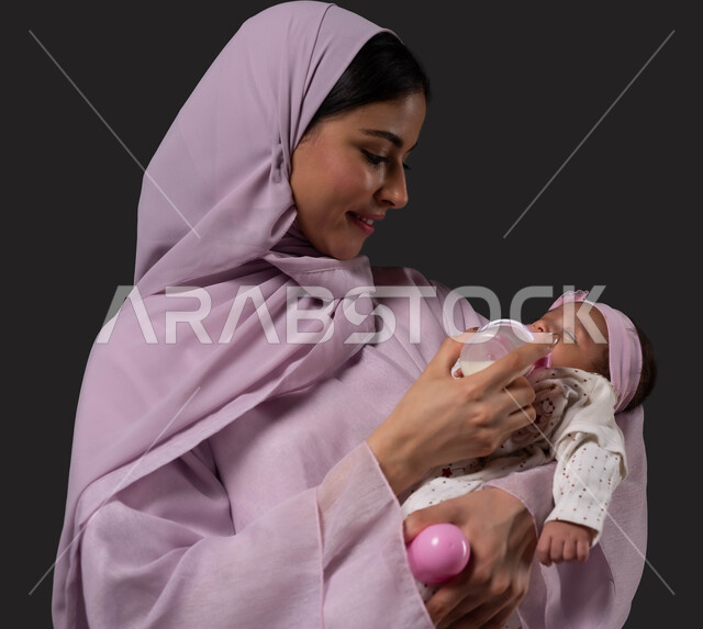 Caring and paying attention to the health and development of children, the concept of motherhood and artificial feeding, a close-up portrait of a smiling Saudi Arabian Gulf mother holding her baby, giving her milk from a feeding bottle, eating healthy food and meals, spending enjoyable times with children, black background