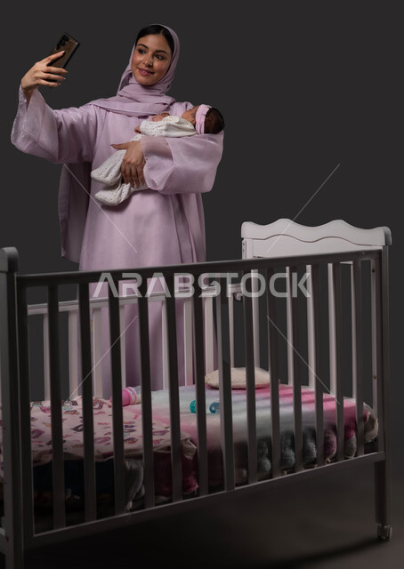 استخدام الأجهزة والتقنيات الحديثة، التقاط صور السليفي التذكارية عبر الهاتف المحمول، بورتريه لأم عربية خليجية سعودية مبتسمة ترتدي العباءة تحمل صغيرتها بحب وحنان، إيماءات الفرح والسرور، توثيق اللحظات السعيدة مع الرضيعة، خلفية سوداء.