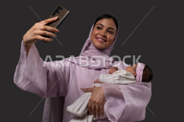 توثيق اللحظات السعيدة مع الرضيعة، التقاط صور السليفي التذكارية عبر الهاتف المحمول، بورتريه لإمرأة عربية خليجية سعودية مبتسمة ترتدي العباءة تحمل صغيرتها بحب وحنان، إيماءات الفرح والسرور، استخدام الأجهزة والتقنيات الحديثة، خلفية سوداء.