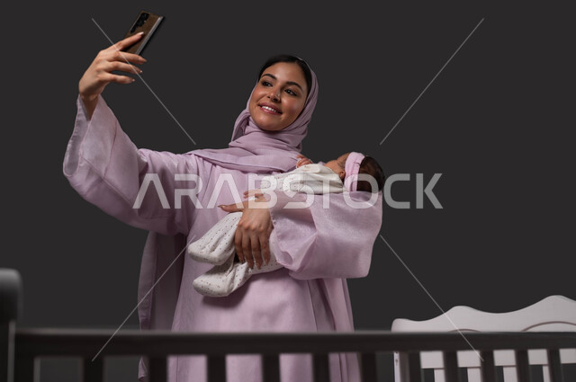 التقاط صور السليفي التذكارية عبر الهاتف المحمول، توثيق اللحظات السعيدة مع الرضيعة، بورتريه لإمرأة عربية خليجية سعودية مبتسمة ترتدي العباءة تحمل صغيرتها بحب وحنان، إيماءات الفرح والسرور، استخدام الأجهزة والتقنيات الحديثة، خلفية سوداء.