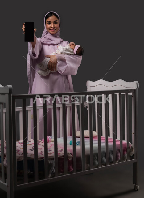استخدام الهاتف المحمول للبحث عن معلومات الأمومة والرعاية الصحية للأطفال، عرض تطبيقات وبرامج العناية بالرضع، بورتريه لإمرأة عربية خليجية سعودية مبتسمة ترتدي العباءة تمسك الجوال بشاشة سوداء فارغة، حمل الرضيعة بإيماءات الحب والحنان، خلفية سوداء.