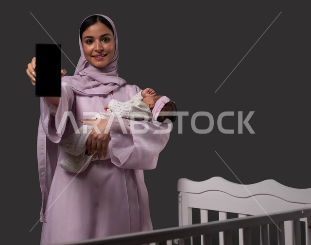 Using audio clips to soothe the infant, online educational courses in parenting and motherhood via mobile phone, portrait of a smiling Saudi Arabian Gulf woman wearing an abaya holding a mobile phone with a blank black screen, holding the baby girl with gestures of love and tenderness, black background