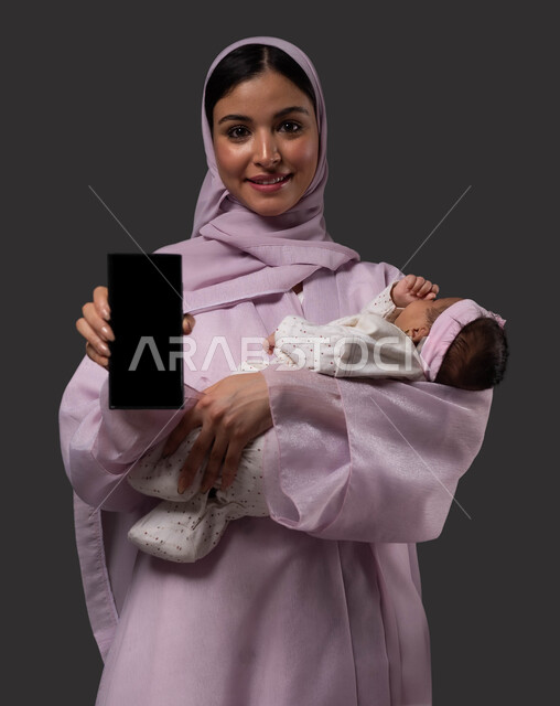 Displaying applications and programs for childcare and care, online educational courses in education and motherhood via mobile phone, a portrait of a smiling Saudi Arabian Gulf woman wearing an abaya holding a mobile phone with a blank black screen, holding the infant with gestures of love and tenderness, a black background