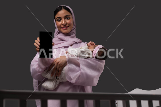 دورات الاونلاين التعليميه في التربيه والامومه عبر الجوال، عرض تطبيقات وبرامج العناية  والرعايه بالاطفال، بورتريه لإمرأة عربية خليجية سعودية مبتسمة ترتدي العباءة تمسك الهاتف المحمول بشاشة سوداء فارغة، حمل الرضيعة بإيماءات الحب والحنان، خلفية سوداء.
