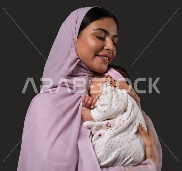 الشعور بالأمان والحب، مفهوم الرعاية والحنان على الأبناء، احتضان الطفلة للتهدئة والاستعماق في النوم، تقوية رابط الأمومة بين الأم ورضيعتها، بورتريه مقرب لإمرأة عربية خليجية سعودية مبتسمة تحمل صغيرتها بعطف وعناية، إيماءات الود والاهتمام، خلفية سوداء.