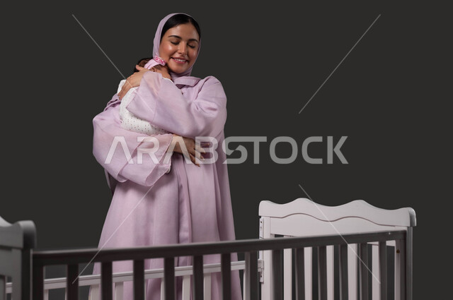 مفهوم الرعاية والحنان على الأبناء، احتضان الطفلة للتهدئة والشعور بالأمان، تقوية رابط الأمومة بين الأم ورضيعتها، بورتريه لإمرأة عربية خليجية سعودية مبتسمة تحمل صغيرتها بعطف وحب، إيماءات الود والاهتمام، خلفية سوداء.
