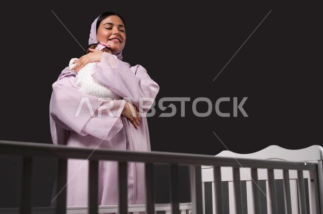 احتضان الطفلة للتهدئة والشعور بالأمان، تقوية رابط الأمومة بين الأم ورضيعتها، بورتريه لإمرأة عربية خليجية سعودية مبتسمة تحمل صغيرتها بعطف وحب، إيماءات الود والاهتمام، مفهوم الرعاية والحنان على الأبناء، خلفية سوداء.