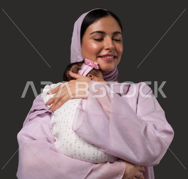 تقوية رابط الأمومة بين الأم ورضيعتها، احتضان الطفلة للتهدئة والشعور بالأمان، بورتريه مقرب لإمرأة عربية خليجية مبتسمة سعودية تحمل صغيرتها بعطف وحب، إيماءات الود والاهتمام، مفهوم الرعاية والحنان على الأبناء، خلفية سوداء.
