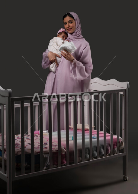 الاستعداد لتغيير البامبرز في السرير، تحمل المسؤولية والعناية بالأطفال، تبديل الحفاظة والمحافظة على نظافة الرضيعة، بورتريه لإمرأة عربية خليجية سعودية مبتسمة تنظر إلى الكاميرا تحمل صغيرتها تحضنها بعطف وحب، خلفية سوداء.