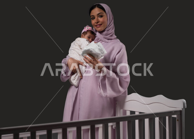 تحمل المسؤولية والعناية بالأطفال، تبديل الحفاظة والمحافظة على نظافة الرضيعة، بورتريه لأم عربية خليجية سعودية مبتسمة تنظر إلى الكاميرا تحمل صغيرتها تحضنها بعطف وحب وحنان، الاستعداد لتغيير البامبرز في السرير، خلفية سوداء.