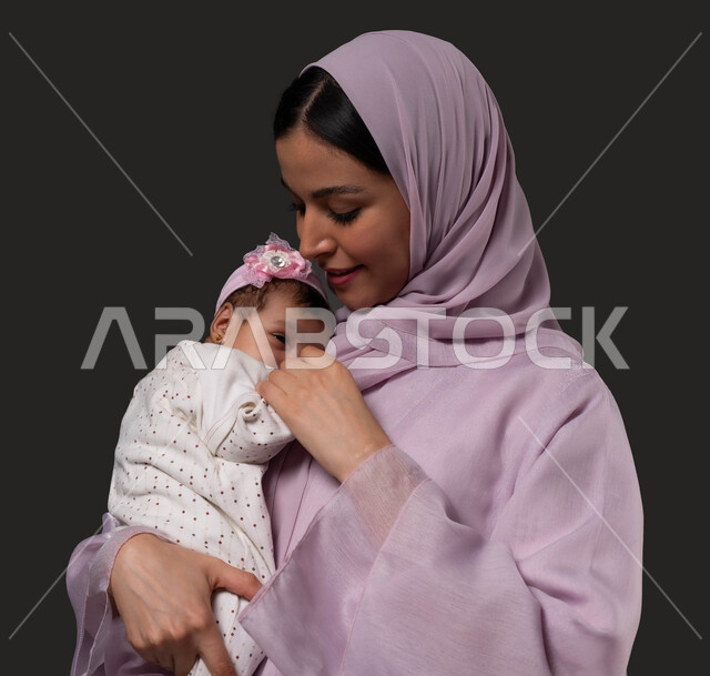 قضاء وقت ممتع مع البنات، حنان الأمهات و تحمل المسؤولية والعناية بالأطفال، بورتريه مقرب لأم عربية خليجية سعودية مبتسمة تحمل طفلتها الرضيعة تمسك يدها وتحضنها بعطف وحب، مشاعر الأمومة والطفولة، خلفية سوداء.