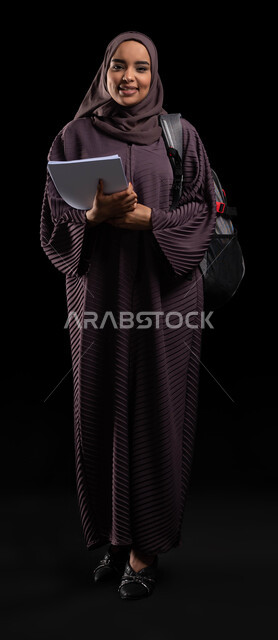 الأكاديميات التعليمية في دول الخليج, الاستعداد للعام الدراسي الجديد, التعليم والتعلم في السعودية, بورتريه لطالبة جامعية عربية خليجية سعودية مبتسمة محجبة ترتدي حقيبة الظهر تمسك الكتب الجامعية وتنظر إلى الكاميرا, صورة الجسم بالطول الكامل, خلفية سوداء