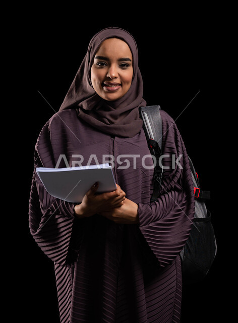 Quality of teaching and learning in Saudi Arabia, educational academies in the Gulf countries, preparing for the new academic year, close-up portrait of a smiling, veiled Saudi Gulf Arab university student wearing a backpack holding university books, black background