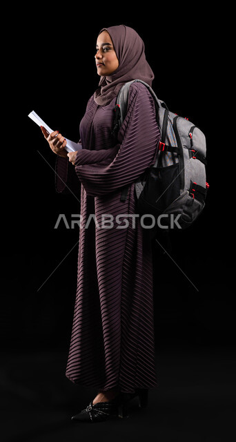 الاستعداد للعام الجامعي الجديد, النظر إلى شيء ما والوقوف بإيماءات الفخر, الأكاديميات التعليمية في دول الخليج, بورتريه من الجانب لطالبة جامعية عربية خليجية سعودية محجبة ترتدي حقيبة الظهر تمسك الكتب الجامعية, صورة الجسم بالطول الكامل, خلفية سوداء