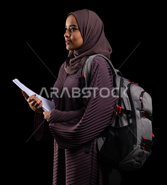 التعلم والتعليم في السعودية, النظر إلى شيء ما والوقوف بإيماءات الفخر, الأكاديميات التعليمية في دول الخليج, الاستعداد للعام الدراسي الجديد, بورتريه من الجانب لطالبة جامعية عربية خليجية سعودية محجبة ترتدي حقيبة الظهر تمسك الكتب الجامعية, خلفية سوداء