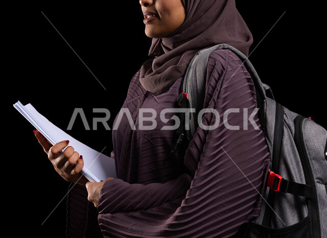 النظر إلى شيء ما والوقوف بإيماءات الفخر, الأكاديميات التعليمية في دول الخليج, الاستعداد للعام الدراسي الجديد, بورتريه مقرب من الجانب لطالبة جامعية عربية خليجية سعودية محجبة مبتسمة ترتدي حقيبة الظهر تمسك الكتب الجامعية, خلفية سوداء