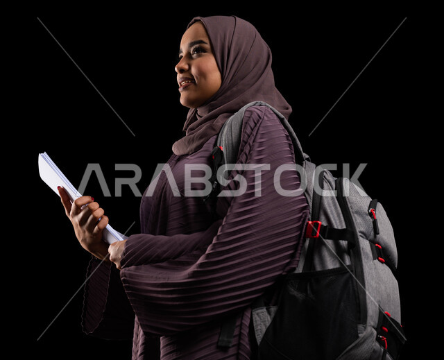 الوقوف بفخر والنظر إلى شيء ما, الأكاديميات التعليمية في دول الخليج, الاستعداد للعام الجامعي الجديد, التعليم والتعلم في السعودية, بورتريه مقرب من الجانب لطالبة جامعية عربية خليجية سعودية محجبة ترتدي حقيبة الظهر تمسك الكتب الجامعية, خلفية سوداء