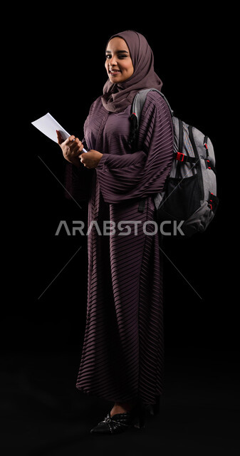 الأكاديميات التعليمية في دول الخليج, الاستعداد للعام الجامعي الجديد, التعليم والتعلم في السعودية, بورتريه لطالبة جامعية عربية خليجية سعودية محجبة ترتدي حقيبة الظهر تمسك الكتب الجامعية وتنظر إلى الكاميرا, صورة الجسم بالطول الكامل, خلفية سوداء