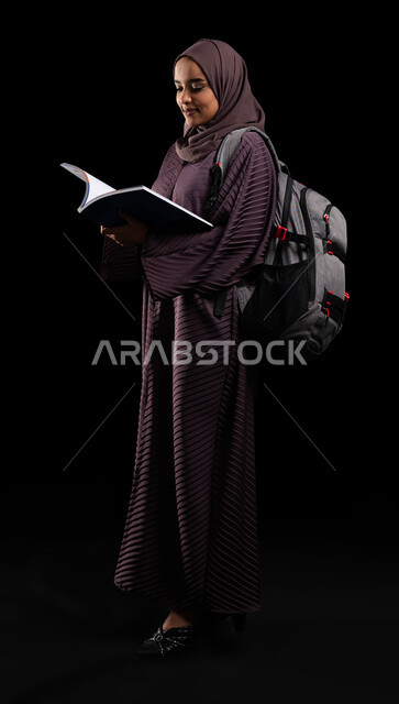 Teaching and learning in Saudi colleges, gestures of walking and going to university, close-up portrait of a veiled Saudi Gulf Arab university student wearing a backpack, tickets with a university book, educational academies in the Gulf countries, full-length body photo, black background