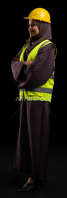 الإعمار والمشاريع الهندسية, بورتريه لمهندسة عربية خليجية سعودية مبتسمة ترتدي سترة الحماية والخوذة تقف بأيدي متقاطعة وتنظر إلى شيء ما, العمل في القطاع الهندسي, تحقيق حلم النجاح والوصول لمهنة الهندسة بإيماءات الفخر, صورة الجسم بالطول الكامل, خلفية سوداء