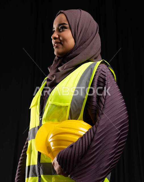 الوقوف والنظر إلى شيء ما, بورتريه مقرب لمهندسة عربية خليجية سعودية مبتسمة ترتدي سترة العمل تمسك خوذة الحماية وتقف بإيماءات الثقة والفخر, العمل والشغف للوصول إلى النجاح, تطور وتنمية السعودية بمجال الهندسة والبناء على يد أبناء الوطن, خلفية سوداء