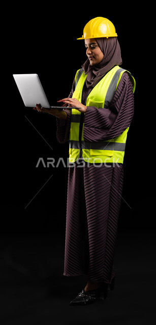 دراسة أساسيات المشروع عبر التطبيقات والتقنيات المتطورة, انجاز مهام العمل عبر الجهاز المحمول, بورتريه لمهندسة عربية خليجية سعودية مبتسمة ترتدي سترة الحماية والخوذة تمسك الابتوب, ادارة المشاريع الهندسية, صورة الجسم بالطول الكامل, خلفية سوداء