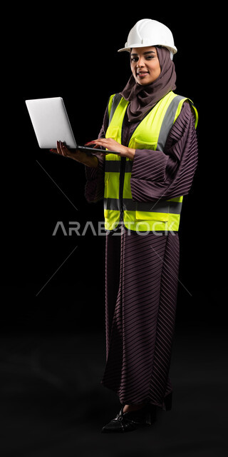 Engineering plans and designs, passion for the engineering profession, use of modern technical devices in the engineering field, portrait of a smiling Saudi Arabian Gulf engineer wearing a protection jacket and helmet, looking at the camera and holding the computer, full-length body portrait, black background