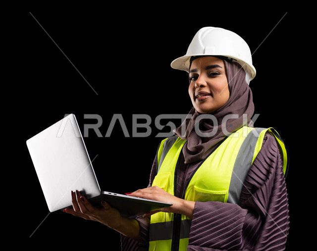 استخدام الأجهزة التقينة الحديثة في المجال الهندسي, بورتريه مقرب لمهندسة عربية خليجية سعودية مبتسمة ترتدي سترة الحماية والخوذة تنظر إلى الكاميرا وتمسك جهاز الحاسوب, تصاميم ومخططات هندسية, مهن ووظائف نسائية, الشغف في مهنة الهندسة, خلفية سوداء