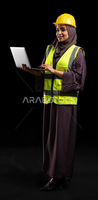 دمج العمل مع التكنولوجيا, دراسة أساسيات المشروع عبر التطبيقات والتقنيات الحديثة, إدارة المشاريع الهندسية, بورتريه لمهندسة عربية خليجية سعودية مبتسمة ترتدي الخوذة وسترة الحماية تمسك بيدها جهاز الحاسوب, صورة الجسم بالطول الكامل, خلفية سوداء