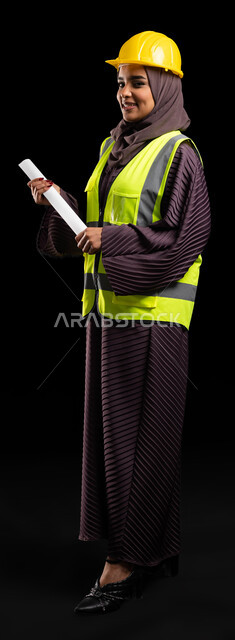 المخططات الانشائية المعمارية, تنمية وتطور السعودية على يد أبناء الوطن, متابعة الأعمال الهندسية, بورتريه لمهندسة عربية خليجية سعودية مبتسمة ترتدي خوذه الحماية وسترة العمل تقف بإيماءات الثقة وتنظر إلى الكاميرا, صورة  الجسم بالطول الكامل, خلفية سوداء