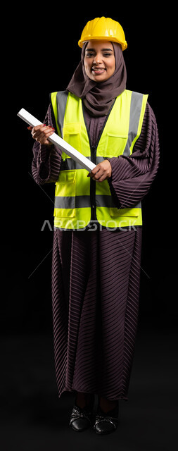 Engineering projects and reconstruction plans, standing confidently and looking at the camera, close-up portrait of a smiling Saudi Arabian Gulf girl wearing a protection vest and helmet holding the construction plan, achieving the dream of success and reaching the engineering profession with proud gestures, full-length body portrait, black background