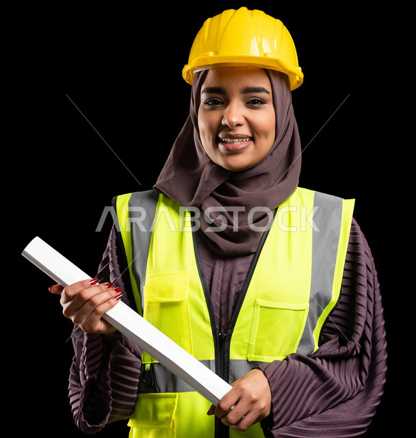 النظر إلى الكاميرا والوقوف بثقة, بورتريه مقرب لفتاة عربية خليجية سعودية مبتسمة ترتدي سترة الحماية والخوذة تمسك مخطط الانشاء, العمل في القطاع الهندسي, تحقيق حلم النجاح والوصول لمهنة الهندسة بإيماءات الفخر, المشاريع الهندسية ومخططات الإعمار, خلفية سوداء