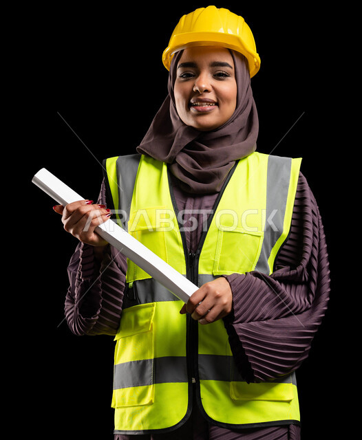 الوقوف بثقة والنظر إلى الكاميرا, بورتريه مقرب لفتاة عربية خليجية سعودية مبتسمة ترتدي سترة الحماية والخوذة تمسك مخطط الانشاء, العمل في القطاع الهندسي, تحقيق حلم النجاح والوصول لمهنة الهندسة بإيماءات الفخر, المشاريع الهندسية ومخططات الإعمار, خلفية سوداء