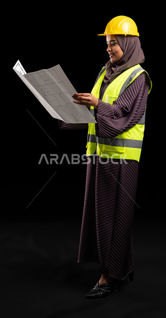 تطور وتقدم السعودية على يد أبناء الوطن, بورتريه لمهندسة عربية خليجية مبتسمة محجبة ترتدي الخوذة وسترة الحماية تمسك مخطط الإعمار, الهندسة والانشاءات المعمارية, الاهتمام بشؤون العمل, تدقيق مخطط الإنشاء, صورة الجسم بالطول الكامل, خلفية سوداء