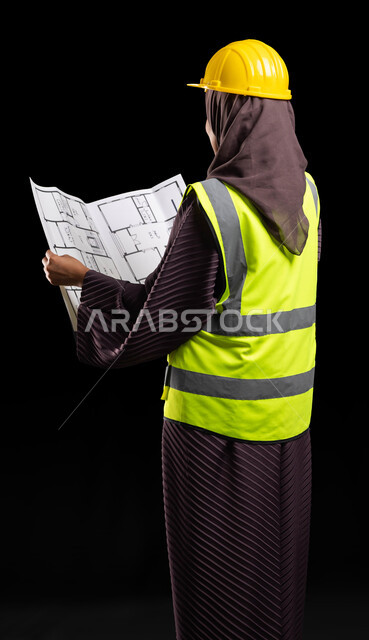 إدارة المشاريع الهندسية, العمل في القطاع الهندسي, التصاميم المعمارية وتدقيق المخططات, بورتريه من الخلف لمهندسة عربية خليجية سعودية محجبة ترتدي سترة الحماية والخوذة تمسك مخطط الإعمار, إدارة المشاريع الهندسية, مهن ووظائف نسائية, خلفية سوداء