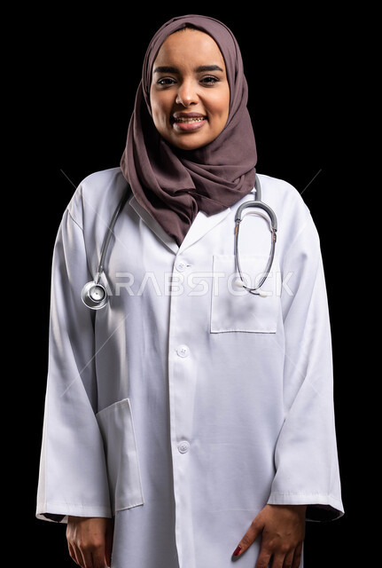 الوقوف بإيماءات الثقة بالنفس والسرور, تقديم المساعده الطبية للمرضى, العمل في المراكز الصحية والمستشفيات, بورتريه مقرب لطبيبة عربية خليجية سعودية مبتسمة ترتدي المعطف الطبي الأبيض وسماعة الفحص تنظر إلى الكاميرا, مفهوم الطب والرعاية الصحية, خلفية سوداء