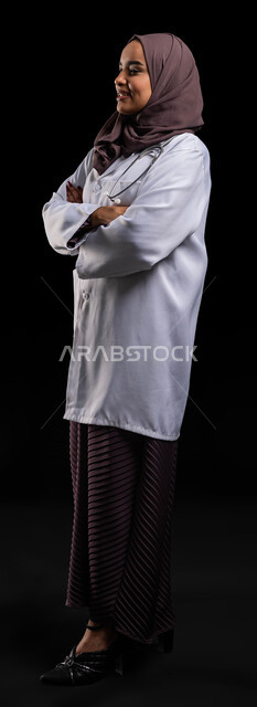 النظر إلى شيء ما والوقوف بثقة, بورتريه من الجانب لطبيبة عربية خليجية سعودية مبتسمة ترتدي المعطف الطبي الأبيض وسماعة الفحص تقف بأيدي متقاطعة, تقديم الخدمات والمساعدة الطبية, العمل في المستشفيات والمراكز الصحية, صورة الجسم بالطول الكامل, خلفية سوداء