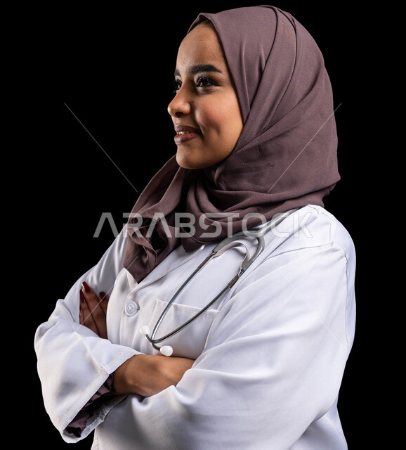 الوقوف بثقة وتحقيق الطموح, بورتريه مقرب لطبيبة عربية خليجية سعودية مبتسمة ترتدي المعطف الطبي الأبيض وسماعة الفحص تقف بأيدي متقاطعة وتنظر إلى شيء ما, تقديم الخدمات والمساعدة الطبية, العمل في المستشفيات والمراكز الصحية, خلفية سوداء