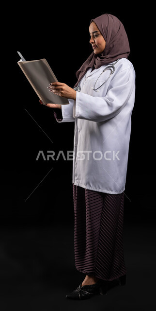 مهن ووظائف سعودية, قراءة نتائج الفحوصات بالسجل الطبي, بورتريه من الجانب لطبيبة عربية خليجية سعودية محجبة ترتدي المعطف الطبي الأبيض تقوم بإعداد التقارير الطبية, تشخيص الحالات المرضية, صورة الجسم بالطول الكامل, خلفية سوداء