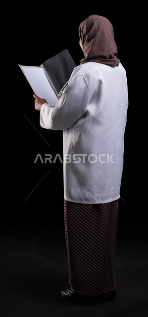 قراءة نتائج الفحوصات بالسجل الطبي, بورتريه من الخلف لطبيبة عربية خليجية سعودية محجبة ترتدي المعطف الطبي الأبيض تقوم بإعداد التقارير الطبية, تشخيص الحالات المرضية, صورة الجسم بالطول الكامل, خلفية سوداء