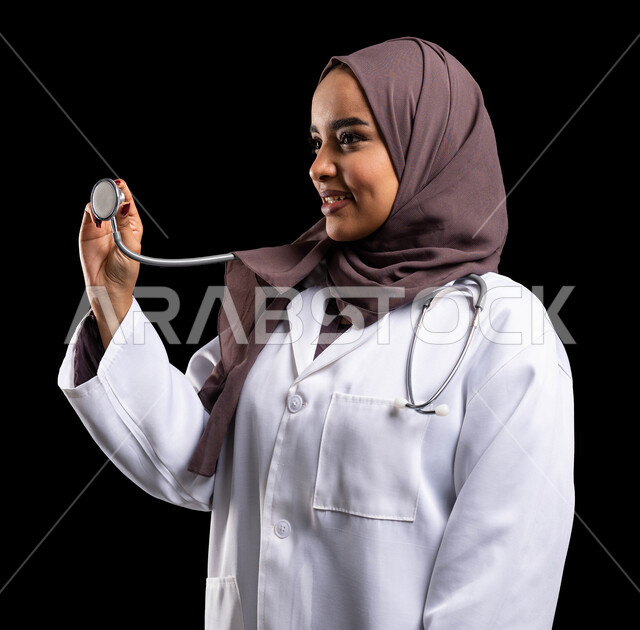 تطوير القطاع الصحي في السعودية، العمل في مجال الرعاية الصحية وتقديم الخدمات الطبية المتميزة، العمل في القطاع الصحي، بورتريه مقرب من الجانب لطبيبة عربية سعودية خليجية محجبة ترتدي المعطف الطبي وتحمل بيدها سماعة الطبيب لسماع نبضات القلب, خلفية سوداء