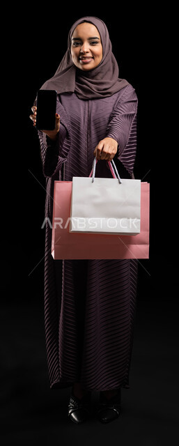 استخدام تطبيقات الهاتف المحمول في شراء المنتجات، التسوق عبر الانترنت من المحلات والمتاجر الالكترونية، بورتريه  لإمرأة عربية خليجية سعودية محجبة ترتدي العباءة تحمل بيدها أكياس تسوق ملونة وهاتف محمول بشاشه سوداء فارغه، صورة الجسم بالطول الكامل، خلفية سوداء.