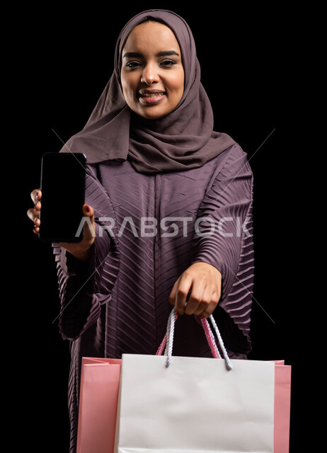 التسوق عبر الانترنت من المحلات والمتاجر الالكترونية، استخدام تطبيقات الهاتف المحمول في شراء المنتجات، بورتريه مقرب لإمرأة عربية خليجية سعودية محجبة ترتدي العباءة تحمل بيدها أكياس تسوق ملونة وهاتف محمول بشاشه سوداء فارغه، خلفية سوداء.