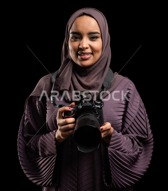 فن توثيق اللحظات، احتراف مهنة التصوير الفوتوغرافي، بورتريه مقرب لإمرأة عربية خليجية سعودية مبتسمة ترتدي عباءة ملونة تمسك بيدها كاميرا تصوير عالية الدقة، التقاط الصور التذكارية، تقنيات التصوير المبتكرة، خلفية سوداء
