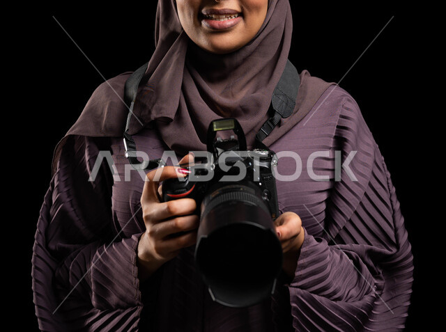 موهبة واحتراف فن التصوير، توثيق الذكريات الثمينة بصور تذكارية، التقاط صور احترافية عالية الدقة، بورتريه مقرب لإمرأة عربية خليجية سعودية محجبة ترتدي العباءة وتحمل بيدها آلة التصوير، استكشاف العالم من خلال العدسة، تقنيات التصوير المبتكرة، خلفية سوداء.