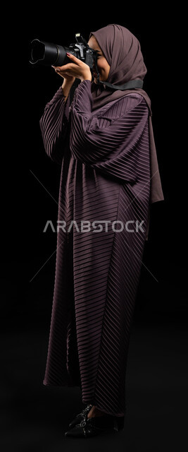 توثيق اللحظات وتخليدها في الذاكرة، كاميرا عالية الدقة، التقاط الصور التذكارية، بورتريه من الجانب لإمرأة عربية خليجية سعودية محجبة ترتدي العباءة وتقوم بالتقاط الصور الفوتوغرافية، استكشاف العالم من خلال العدسة، تقنيات التصوير المبتكرة، صورة الجسم بالطول الكامل، خلفية سوداء.